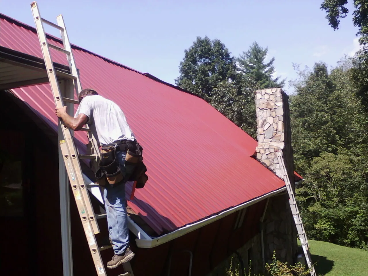 Roofer on a ladder beside a red metal roof and stone chimney — Soffit & Fascia Repair by Roofing Brothers in Joice